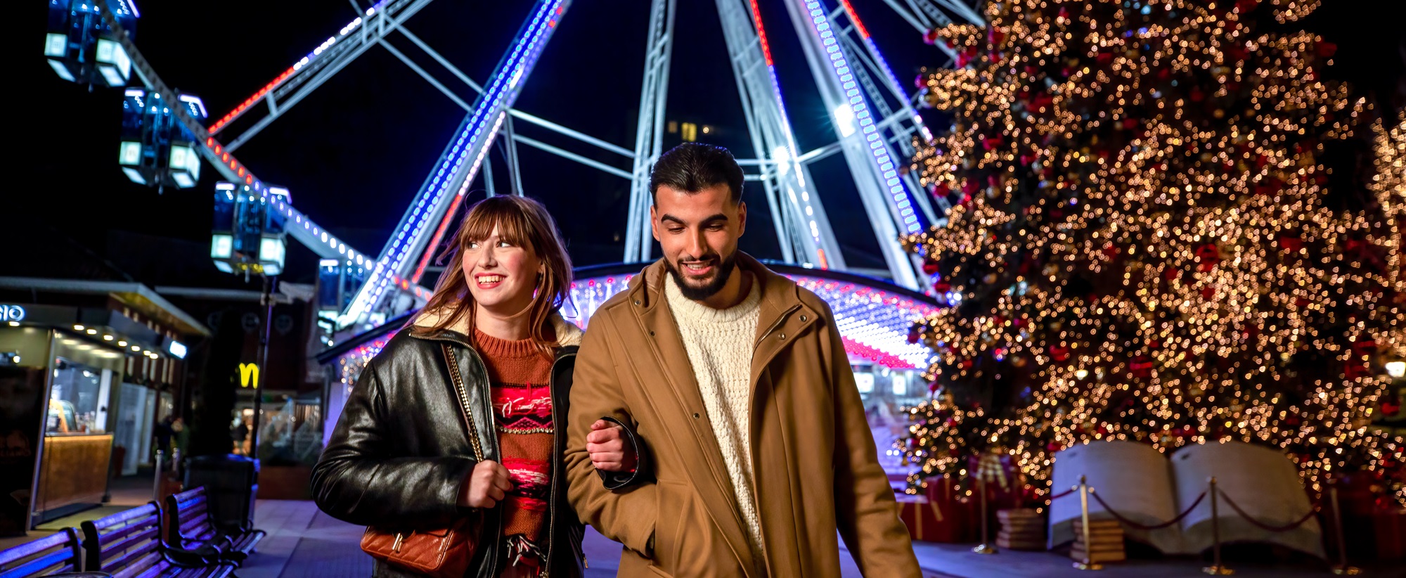 Twee mensen wandelen bij het verlichte reuzenrad en de kerstboom in Designer Outlet Roermond.
