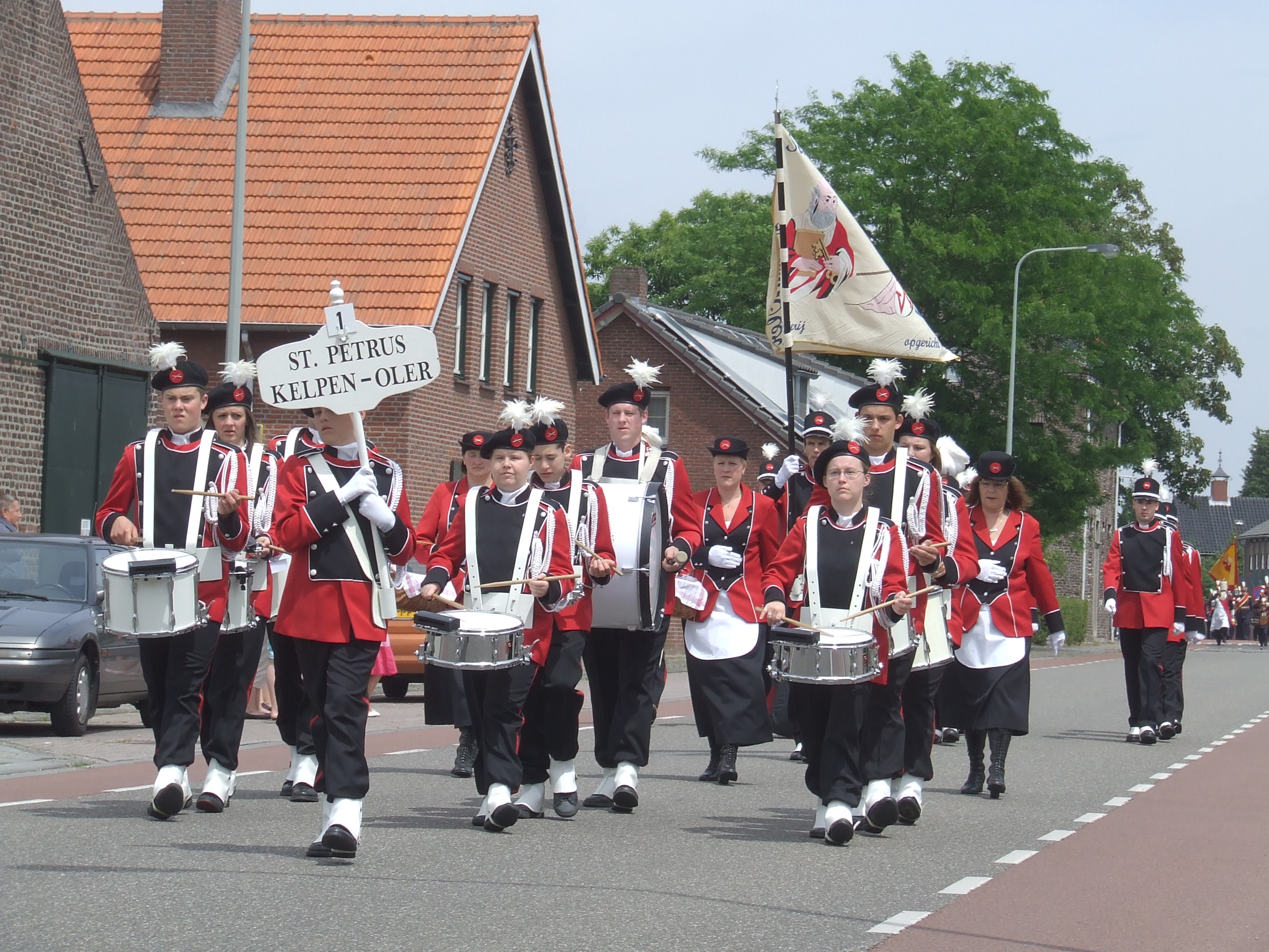 Een schutterij in rood-zwarte uniformen marcheert door een straat tijdens een optocht, met trommelaars en vaandeldragers van St. Petrus Kelpen-Oler