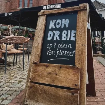 Krijtbord bij De Kompenie in Neer met de tekst "Kom d’r bie op ’t plein van plezeer", voor het terras op een geplaveid dorpsplein met zicht op de kerk.