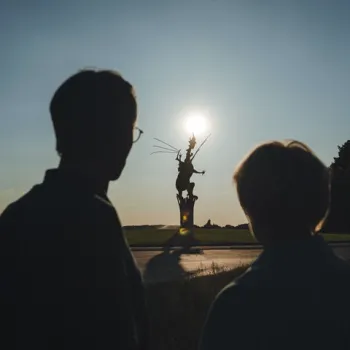 Twee silhouetten van een man en een vrouw kijken bij zonsondergang naar de draak op de rotonde in Beesel, midden in het Limburgse landschap.